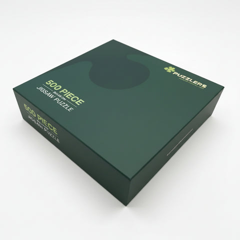 A dark green rectangular box for a 500-piece jigsaw puzzle, photographed at an angle on a clean white background. The box features gold text reading '500 PIECE JIGSAW PUZZLE' and a gold 'PUZZLERS' logo.
